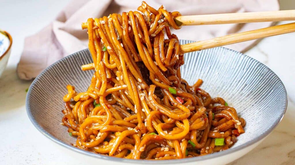 A bowl of stir-fried noodles with sesame seeds and green onions, held with chopsticks.
