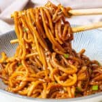 A bowl of stir-fried noodles with sesame seeds and green onions, held with chopsticks.