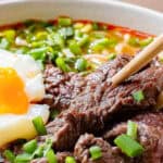 A bowl of noodle soup with beef slices, a soft-boiled egg, and chopped green onions. Chopsticks hold a piece of beef.