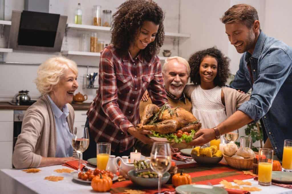 Family gathered around a dining table, sharing a Thanksgiving meal with a roasted turkey as the centerpiece. The table is decorated with candles and pumpkins.