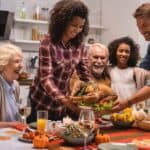 Family gathered around a dining table, sharing a Thanksgiving meal with a roasted turkey as the centerpiece. The table is decorated with candles and pumpkins.