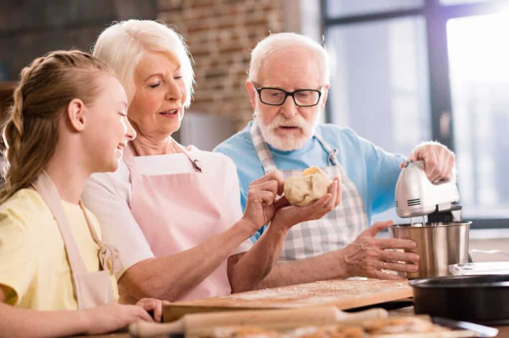 Three people in a kitchen, two older adults and a child, baking together. One holds dough while another uses a stand mixer. They all wear aprons.