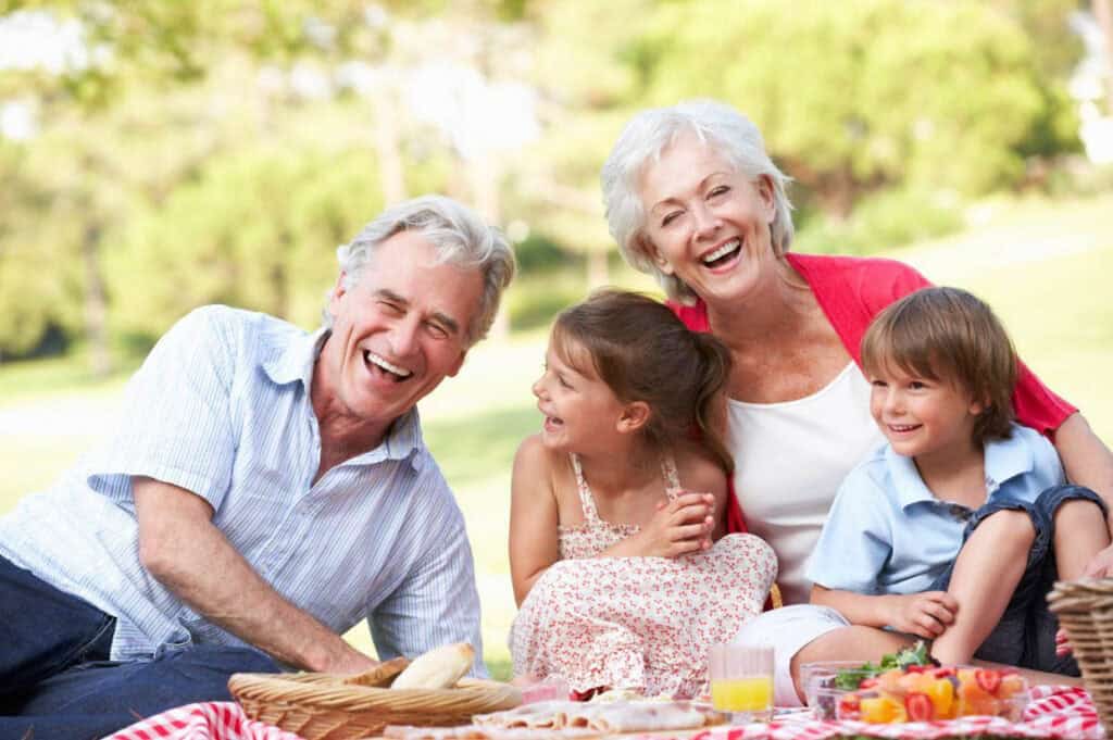 Elderly couple and two children laughing together at a picnic, surrounded by food and greenery.