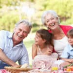 Elderly couple and two children laughing together at a picnic, surrounded by food and greenery.