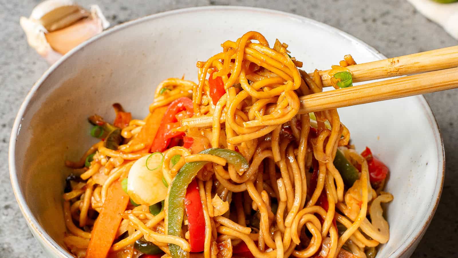 A bowl of stir-fried noodles with colorful vegetables like red and green bell peppers, garnished with spring onions. Chopsticks are lifting a portion of the noodles. A garlic bulb is visible in the background.