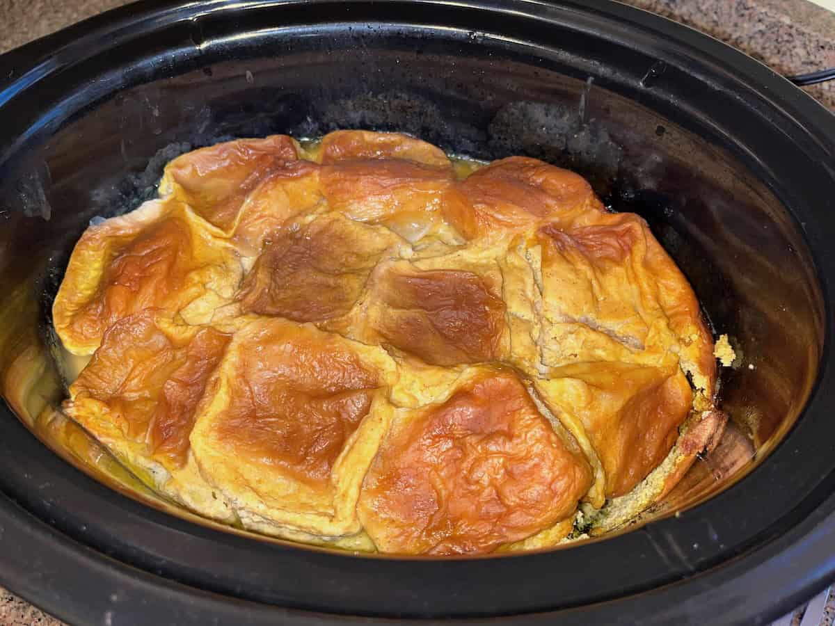 A baked dessert with a golden-brown crust sits in a black slow cooker.