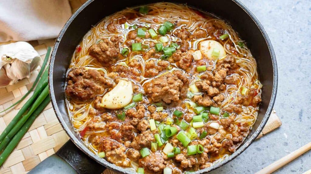 Skillet with noodle soup, ground meat, sliced garlic, and chopped green onions.