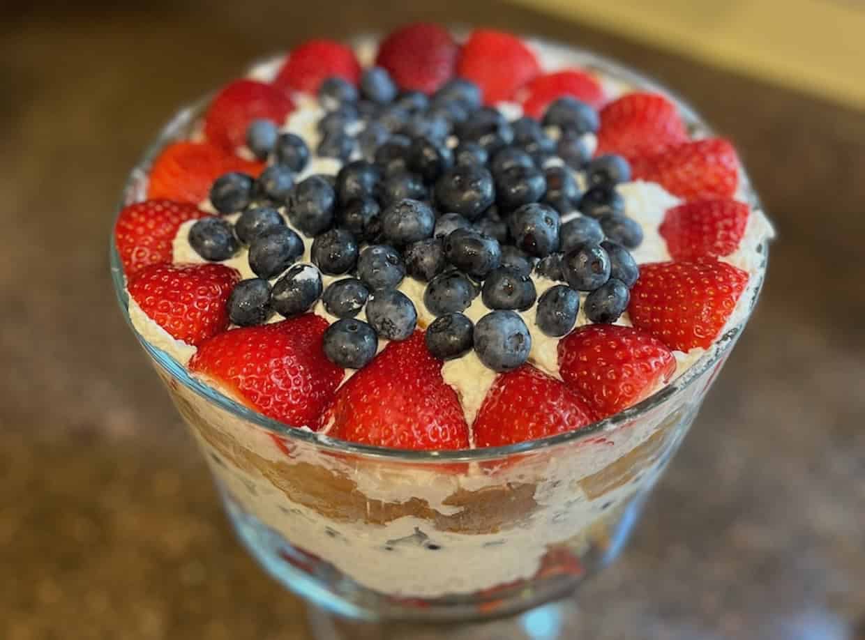 A layered trifle dessert in a glass bowl, topped with whole strawberries and blueberries. The layers include cake, cream, and fruit.