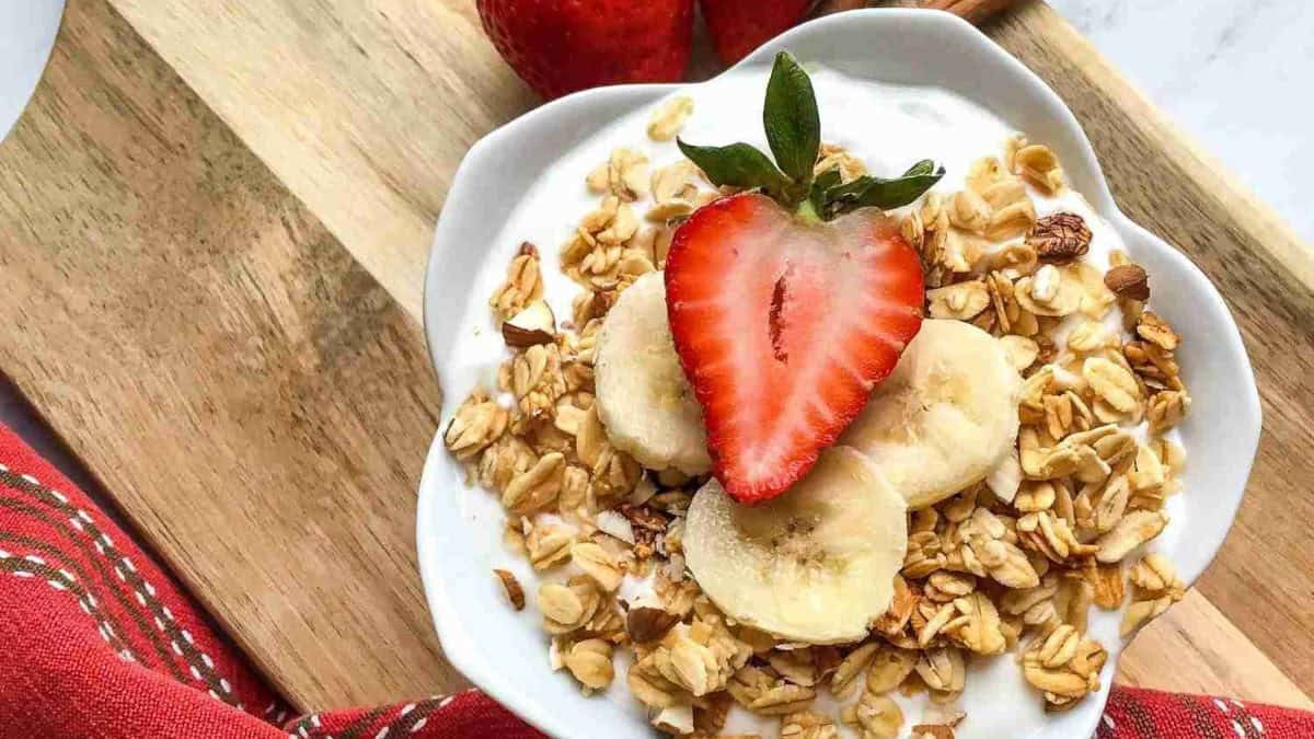 A bowl of yogurt topped with granola, banana slices, and a strawberry on a wooden board with a red cloth.