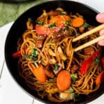 A bowl of stir-fried noodles with beef, carrots, red bell peppers, and greens. Hand using chopsticks to pick up noodles.