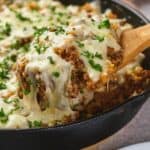 A wooden spoon lifting a serving of italian ground beef skillet garnished with parsley from a cast-iron skillet.