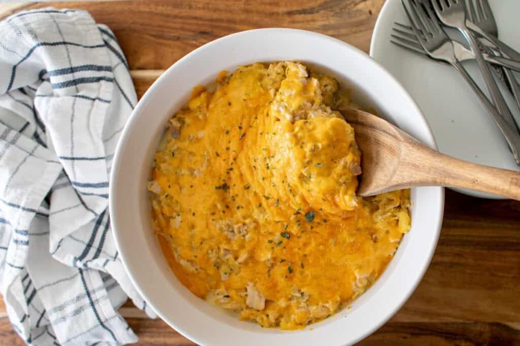 A bowl of cheesy casserole with a wooden spoon, next to a striped cloth and a stack of plates with forks.