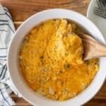 A bowl of cheesy casserole with a wooden spoon, next to a striped cloth and a stack of plates with forks.