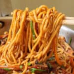 Chopsticks holding a portion of stir-fried noodles with vegetables from a pan.