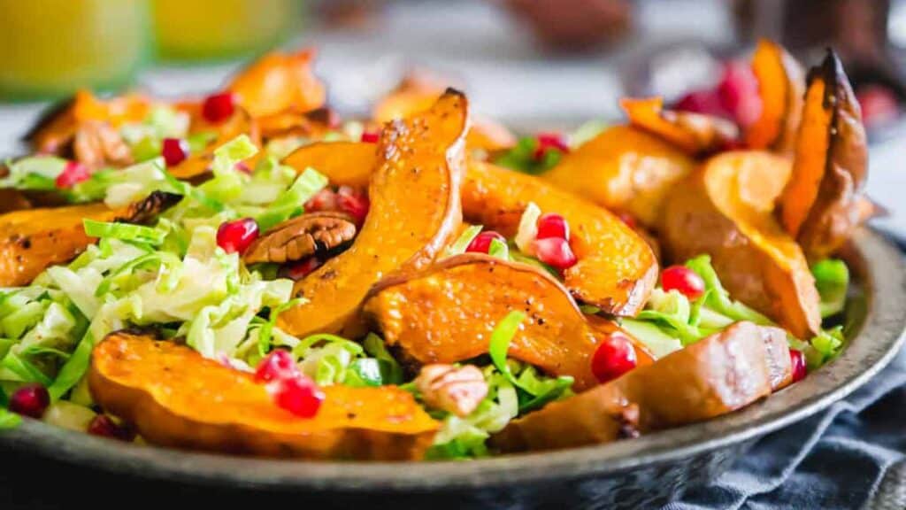 A salad featuring roasted squash slices, shredded greens, pecans, and pomegranate seeds on a plate.