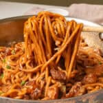 A skillet with cooked noodles and diced meat being stirred with chopsticks.