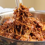 A skillet filled with cooked noodles and chunks of meat, being stirred with tongs.