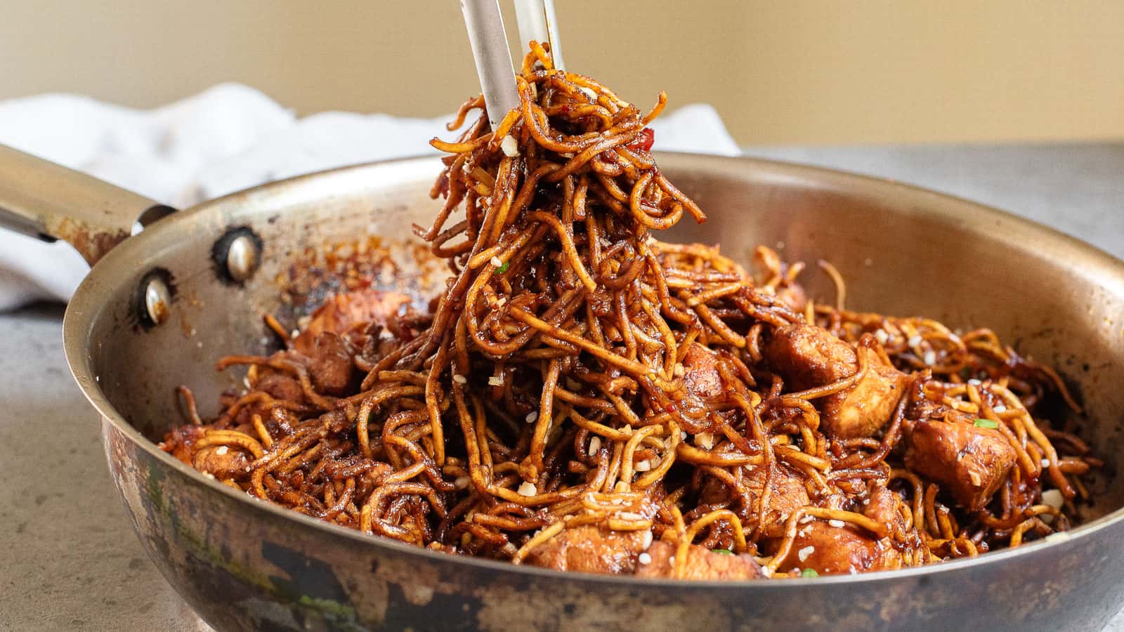 A skillet filled with stir-fried noodles and pieces of chicken being mixed with chopsticks.