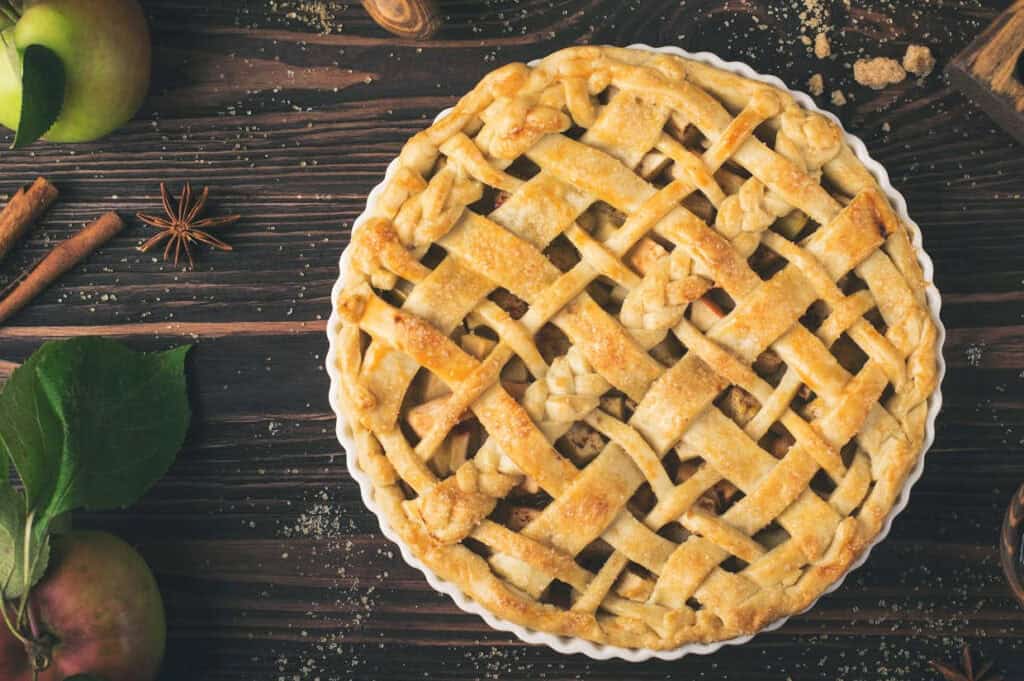 A lattice-topped apple pie in a white dish on a wooden table, surrounded by green apples, cinnamon sticks, and star anise.