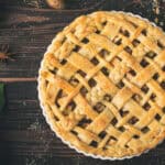 A lattice-topped apple pie in a white dish on a wooden table, surrounded by green apples, cinnamon sticks, and star anise.