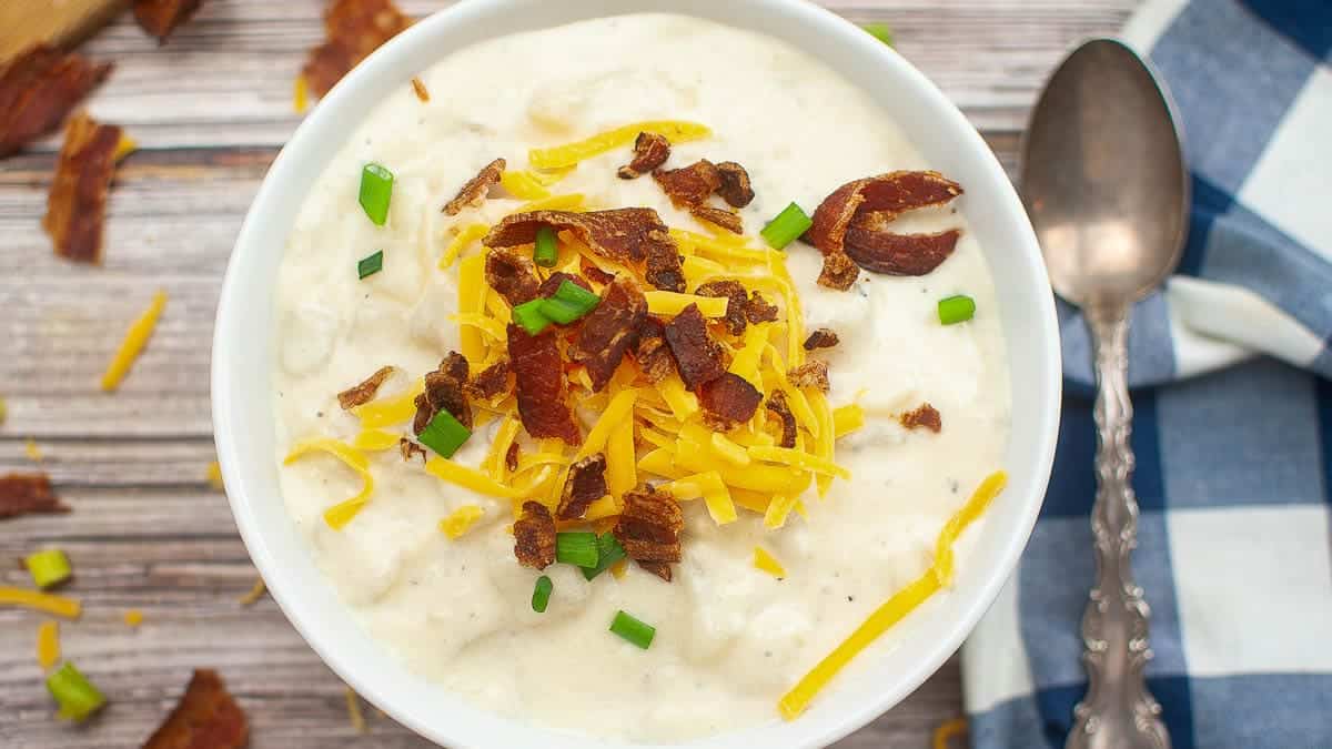 A bowl of creamy potato soup topped with shredded cheddar cheese, chopped green onions, and crumbled bacon, next to a spoon and a blue and white checkered napkin.