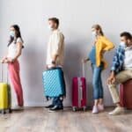 Four people wearing face masks stand in a line with their luggage against a white wall.