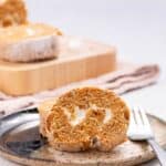 A slice of pumpkin roll cake with cream filling on a plate, fork beside it. More slices and a rolled cake are on a cutting board in the background.