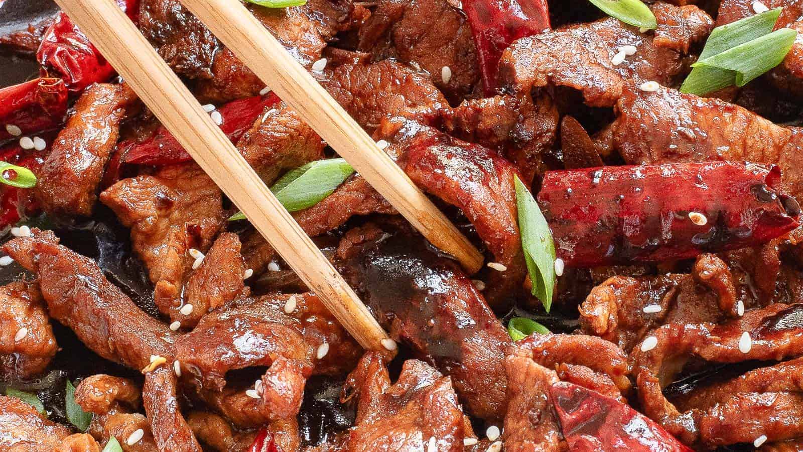 Close-up of a dish featuring sliced beef with red chilies, garnished with sesame seeds and green onions. Chopsticks rest on top.