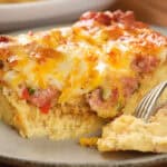 Close-up of a slice of cheesy casserole with bits of meat and vegetables on a plate, with a fork taking a piece.