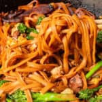 A pan of stir-fried noodles with broccoli, chicken, and egg, being served with chopsticks.