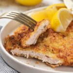 Piece of chicken cutlet on a fork resting over a larger portion with a lemon wedge.