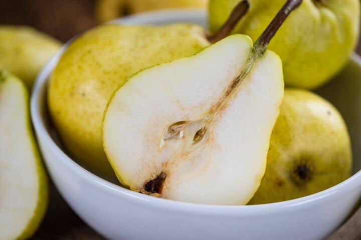 A halved pear with visible seeds rests in a white bowl alongside whole pears.