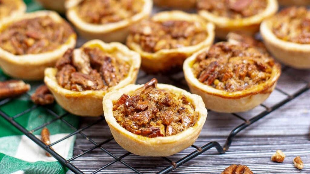 Mini pecan pies cooling on a black wire rack, placed on a wooden surface.