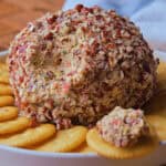 Pineapple cheese ball coated with chopped nuts is placed on a white plate surrounded by round crackers.