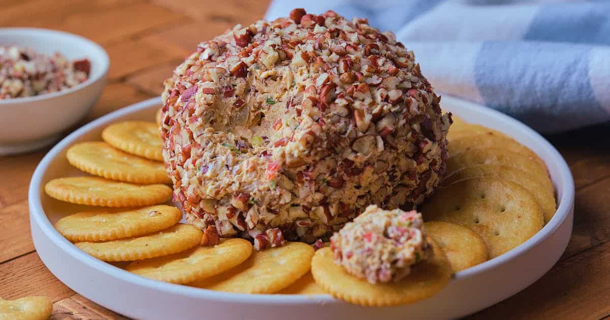 Pineapple cheese ball coated with chopped nuts is placed on a white plate surrounded by round crackers.