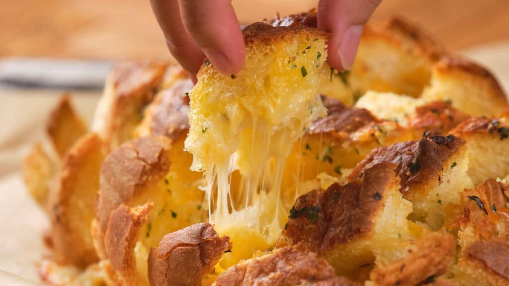 A hand pulls a piece from a cheesy garlic pull-apart bread, with melted cheese stretching.