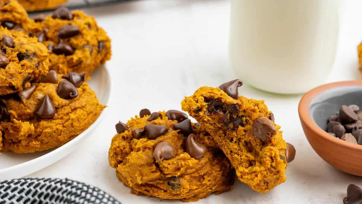 Plate of pumpkin cookies with chocolate chips, alongside a bowl of chocolate chips and a glass of milk on a white surface.