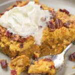 A slice of pumpkin dump cake topped with whipped cream and nuts on a white plate, with a spoon resting beside it.