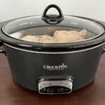 Black Crock-Pot slow cooker on a wooden table with lid closed, cooking food inside.