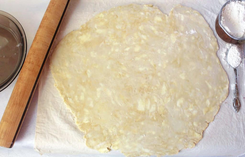 Rolled out pie dough on parchment paper beside a wooden rolling pin and a measuring cup with flour.