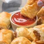 A hand dips a sausage roll into a bowl of ketchup on a plate with several other sausage rolls.