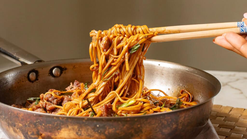 Chopsticks lifting a portion of stir-fried noodles with meat and vegetables from a pan.