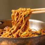 Chopsticks lifting a portion of stir-fried noodles with meat and vegetables from a pan.