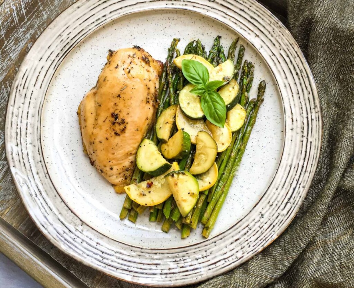 A plate with grilled chicken breast, asparagus, and zucchini slices, garnished with basil, on a wooden table.