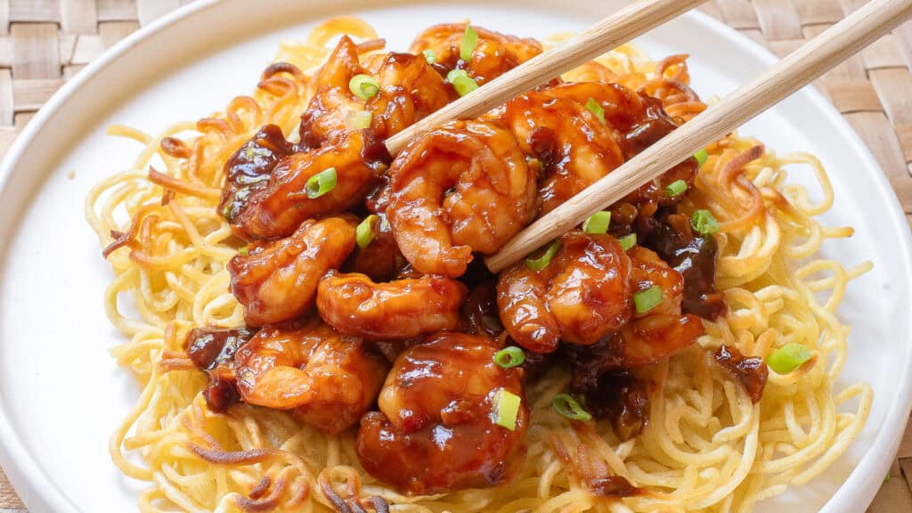 Chopsticks holding honey walnut shrimp on a bed of crispy noodles, garnished with green onions, served on a white plate.