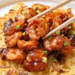 Chopsticks holding honey walnut shrimp on a bed of crispy noodles, garnished with green onions, served on a white plate.