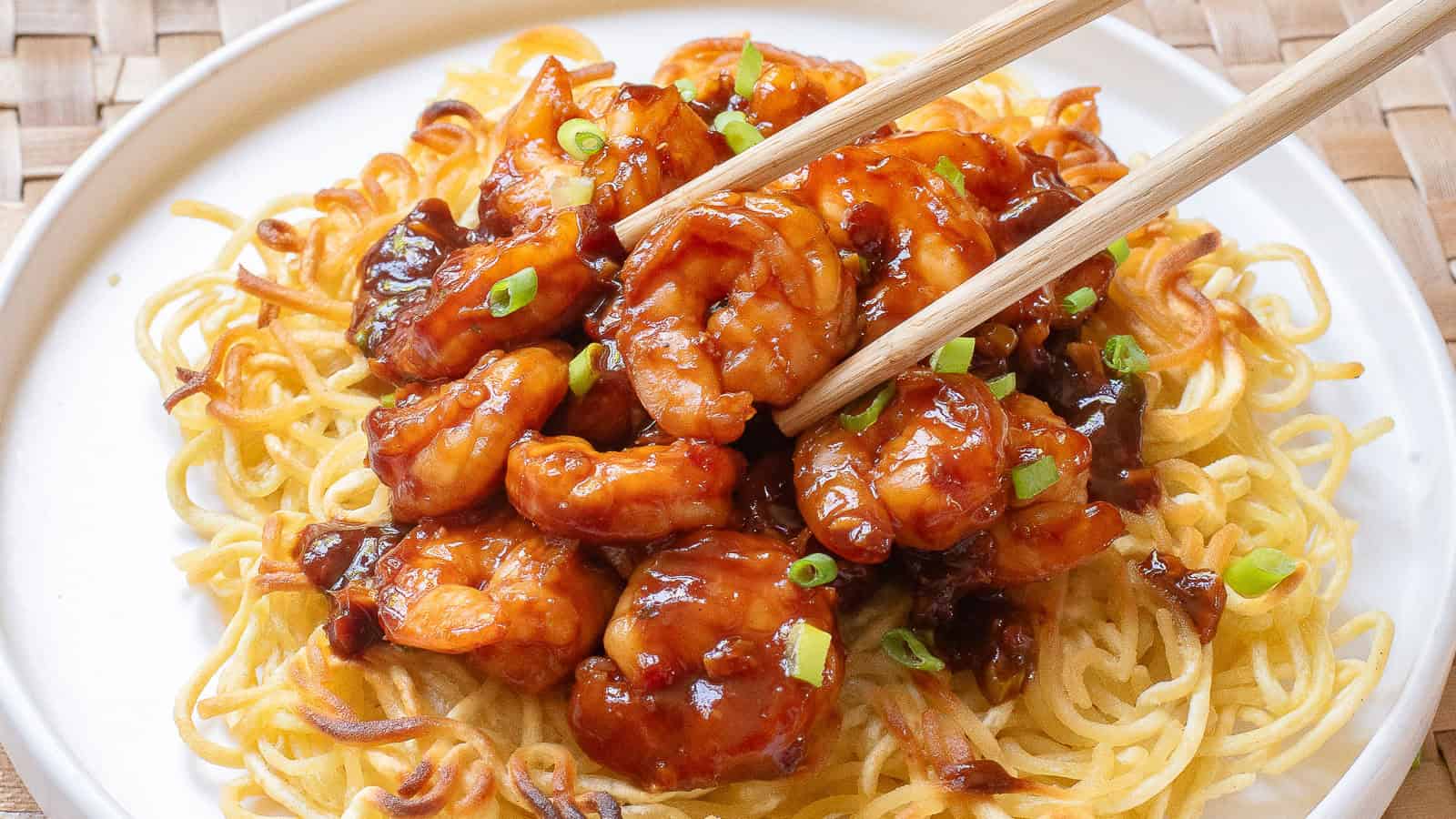 Chopsticks holding a spicy prawn on a bed of crispy noodles, garnished with green onions, served on a white plate.