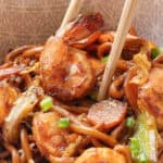 A bowl of shrimp lo mein with noodles, sliced vegetables, and green onions, held with chopsticks.