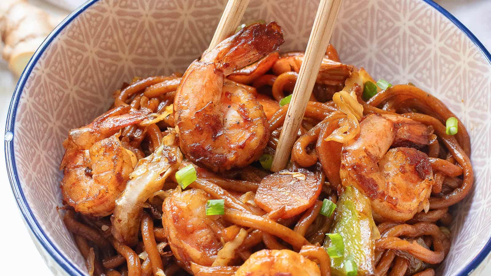 A bowl of shrimp lo mein with noodles, shrimp, and vegetables, topped with green onion slices. Chopsticks are placed on top of the dish. The bowl has a patterned design on the inside rim.