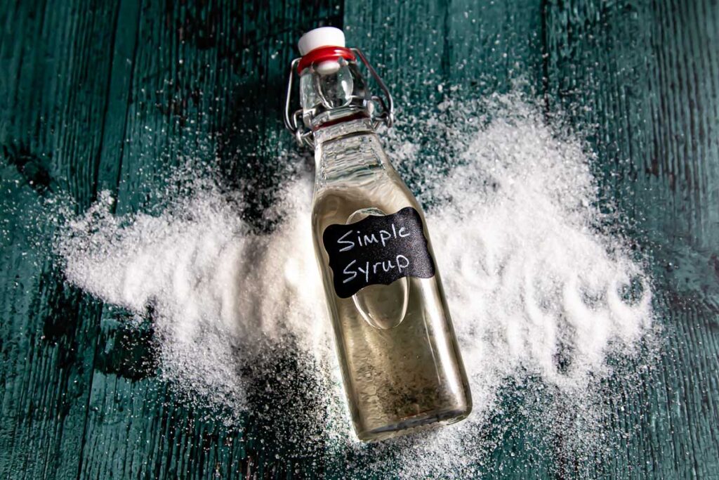 A glass bottle labeled "Simple Syrup" rests on a green surface, surrounded by spilled granulated sugar.